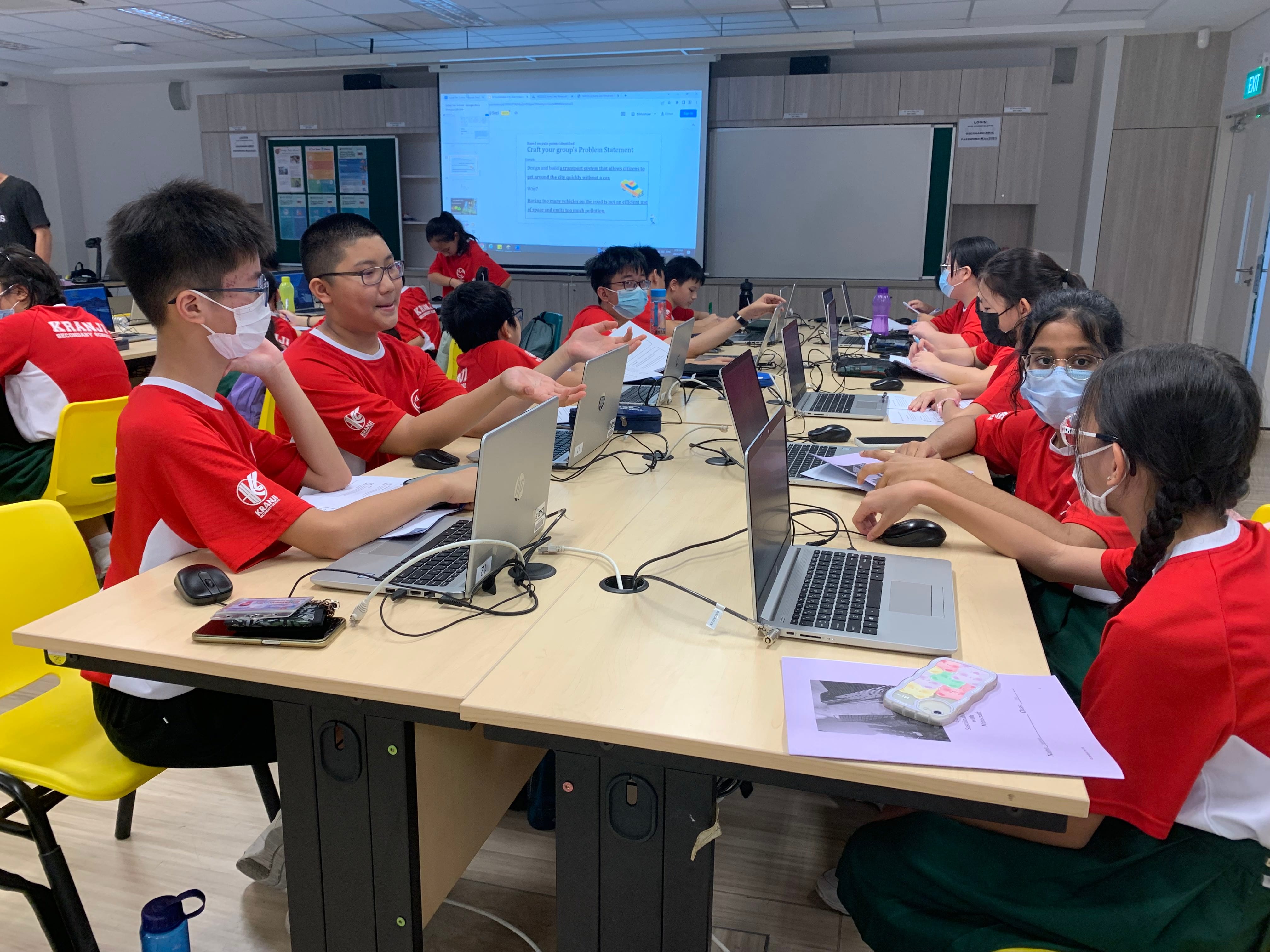 Students in red uniforms and masks work on laptops at classroom tables with a projected presentation visible.